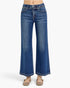A person wearing blue wide-leg denim jeans with a frayed waistband and beige flat sandals, shown from the waist down against a light gray studio background.