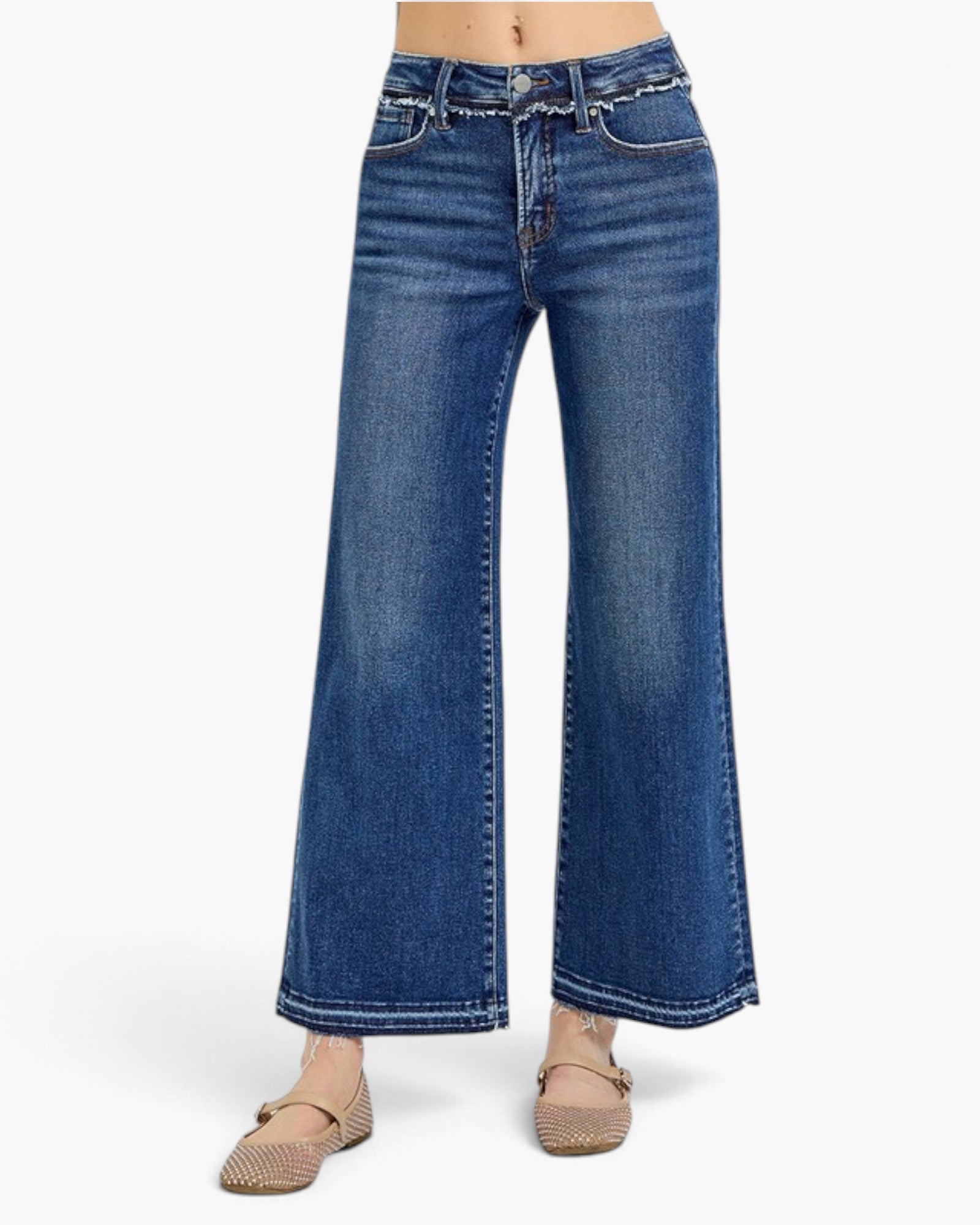 A model is wearing a pair of wide-leg blue denim jeans with decorative silver studs along the waistband, paired with beige flat shoes, shown against a plain white studio background.