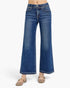 A model is wearing a pair of wide-leg blue denim jeans with decorative silver studs along the waistband, paired with beige flat shoes, shown against a plain white studio background.