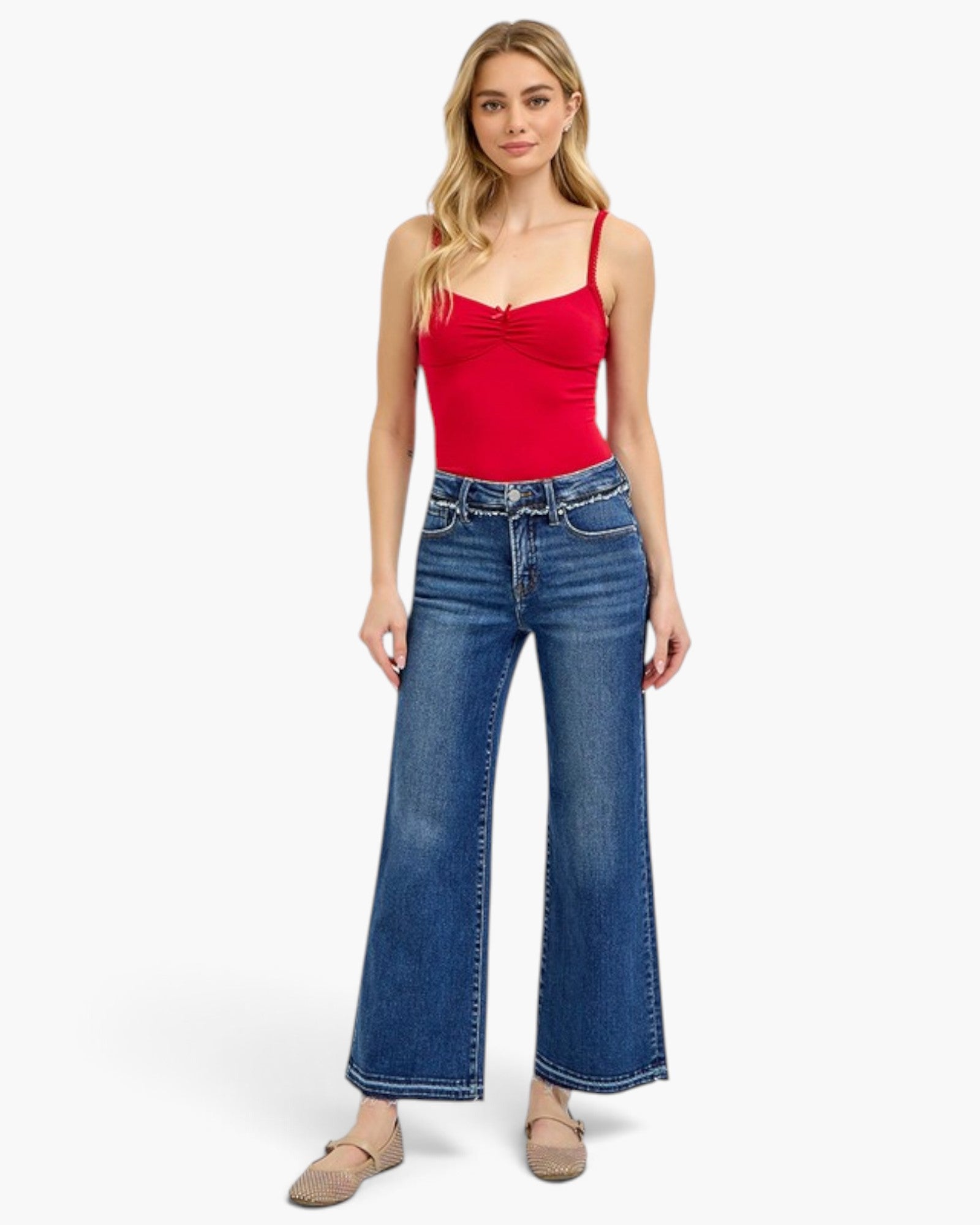 A woman modeling a bright red spaghetti-strap camisole top paired with high-waisted, wide-leg blue denim jeans and beige flat shoes, set against a plain white studio background.