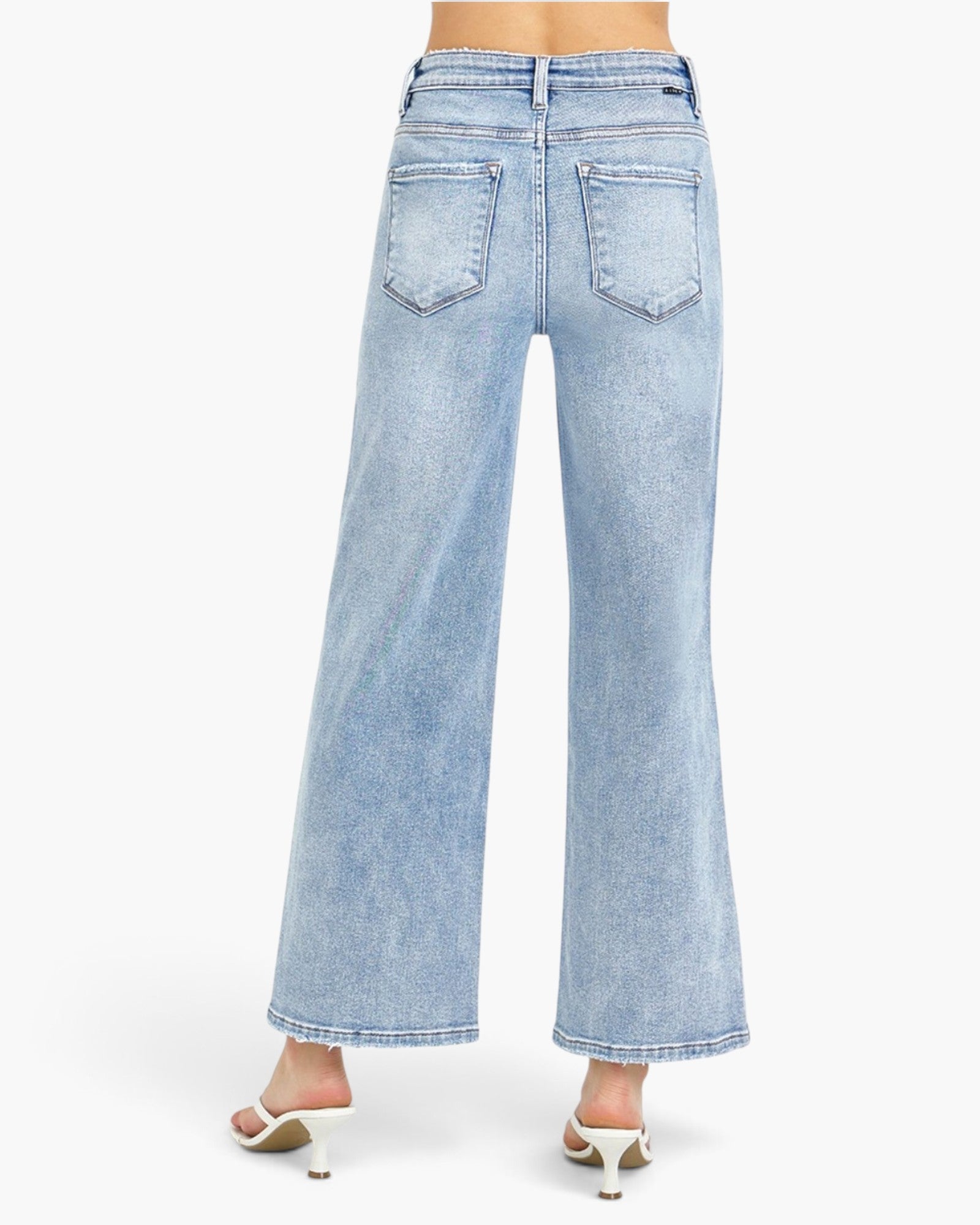 Light wash blue wide-leg denim jeans shown from the back on a model, featuring a high waist, back pockets, and a flared leg, paired with white heeled sandals against a plain white background.