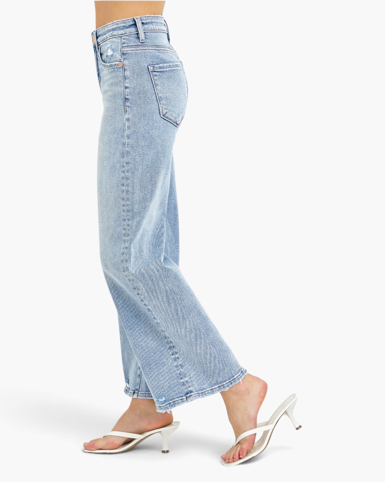 Light wash blue cropped flare jeans shown on a model from the side, paired with white heeled thong sandals against a clean white studio background.