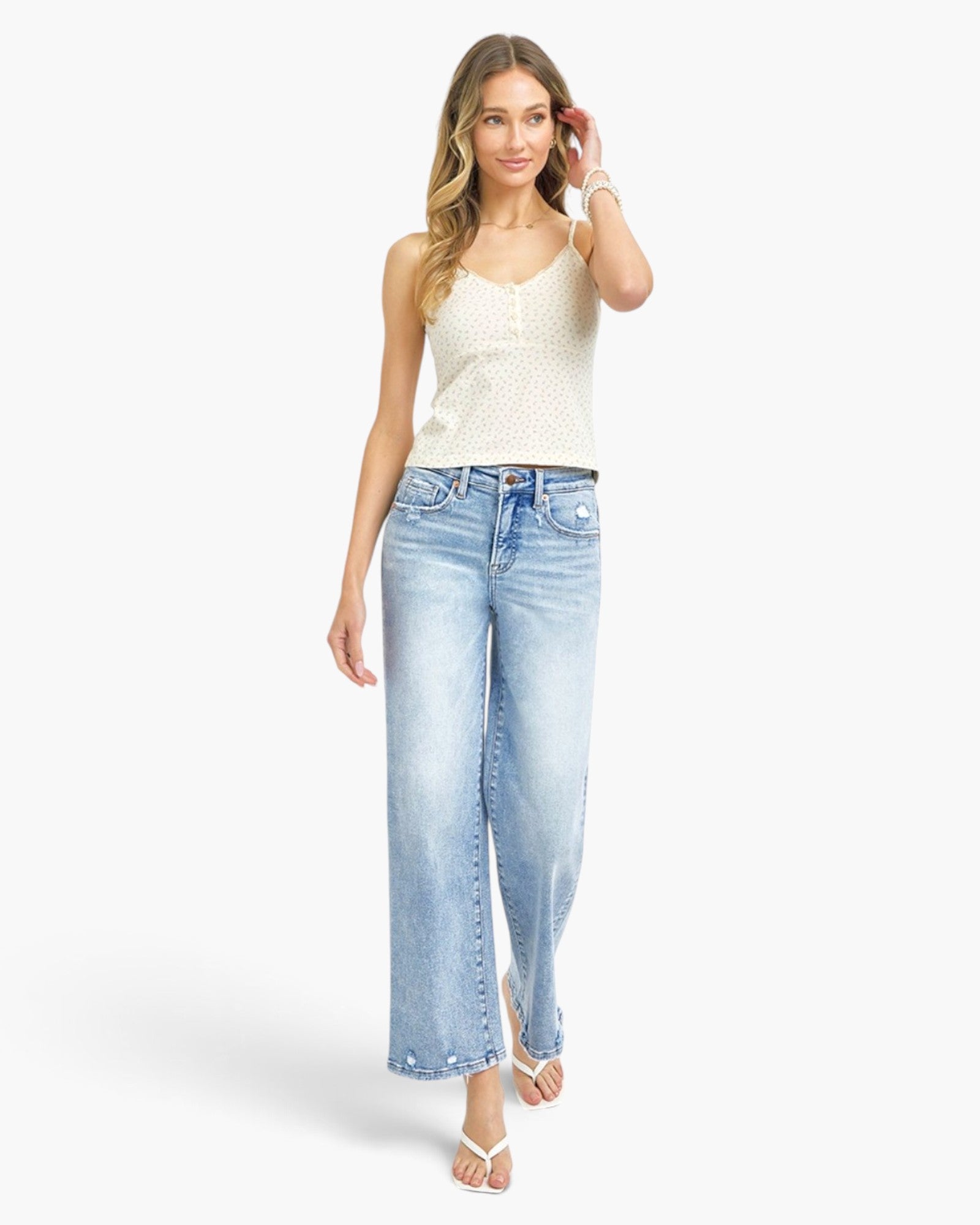 A woman modeling a pair of light-wash wide-leg cropped jeans with a white sleeveless knit top, standing against a plain white studio background.