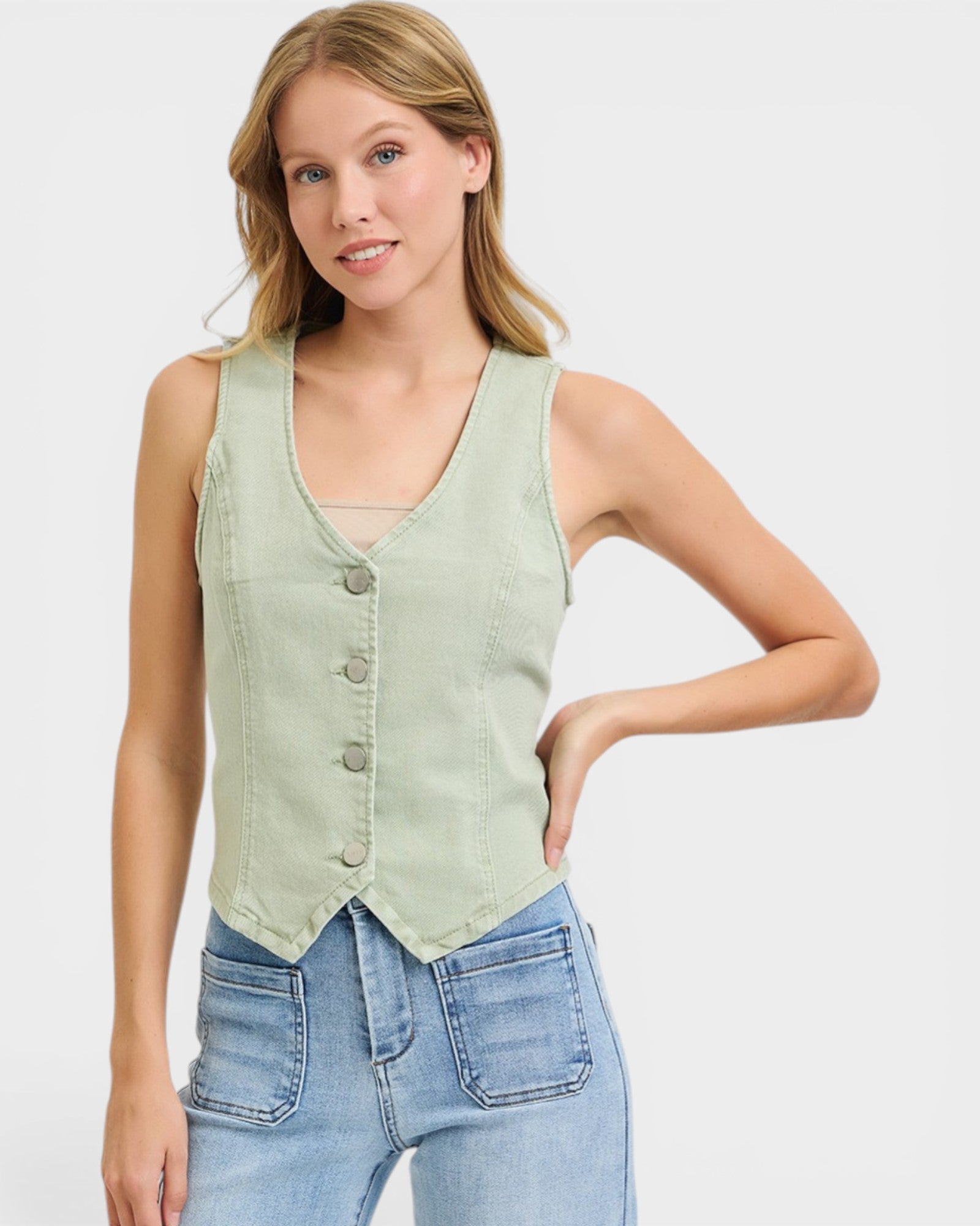 Woman wearing a light green vest over a beige top with blue jeans on a white background