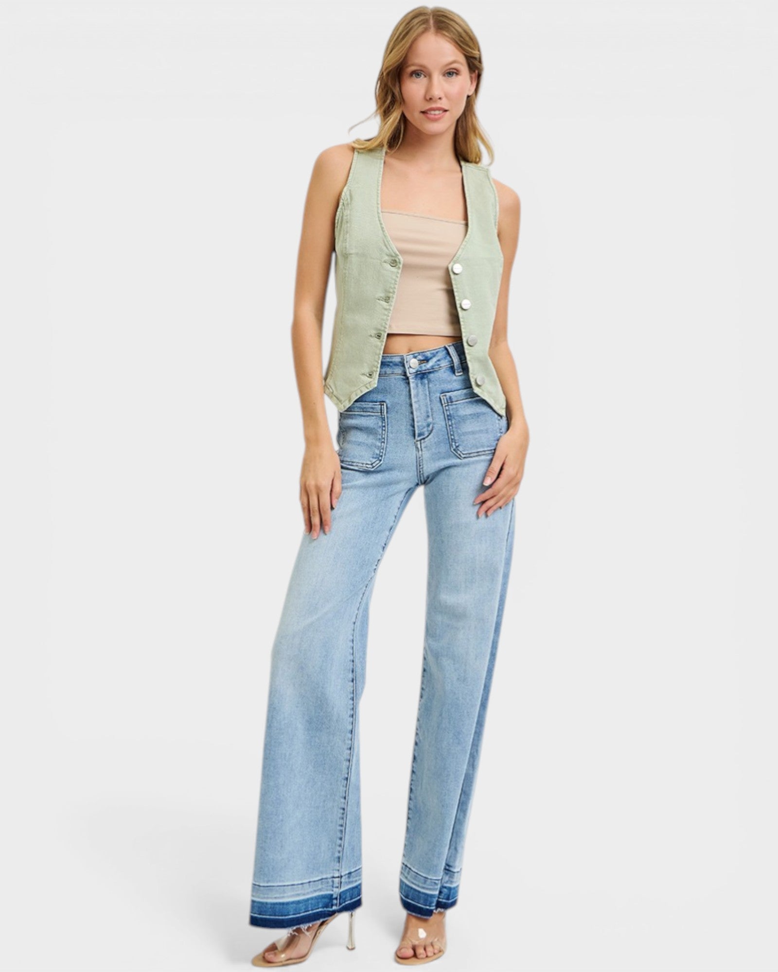 Woman wearing a light green sleeveless jacket, beige top, and blue jeans on a white background