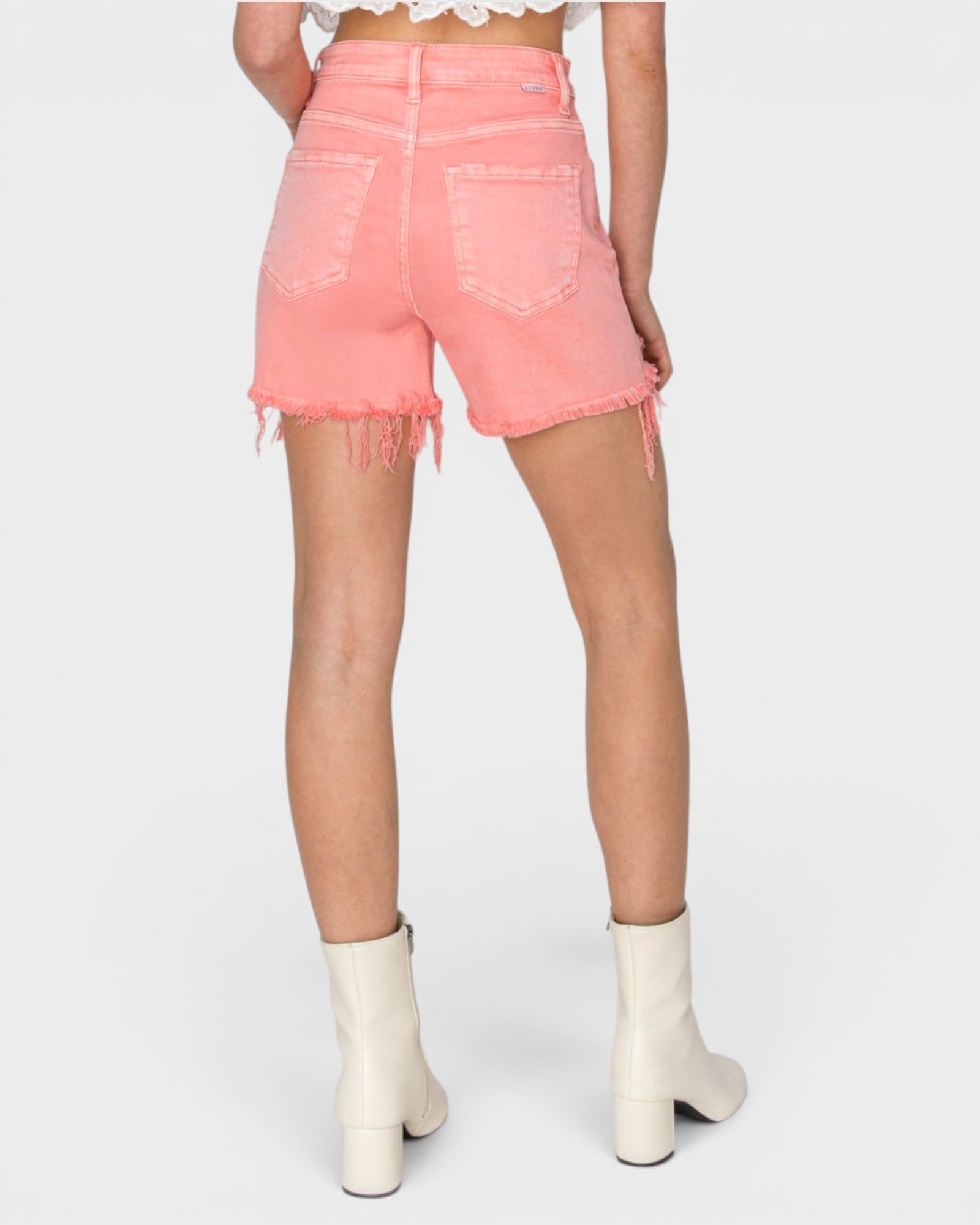 A model shown from the back wearing high-waisted, light pink denim shorts with a frayed hem and white heeled ankle boots, set against a plain light gray background.