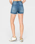 Rear view of a person wearing medium-wash blue denim shorts with a raw, frayed hem and white sneakers, against a light gray studio background.