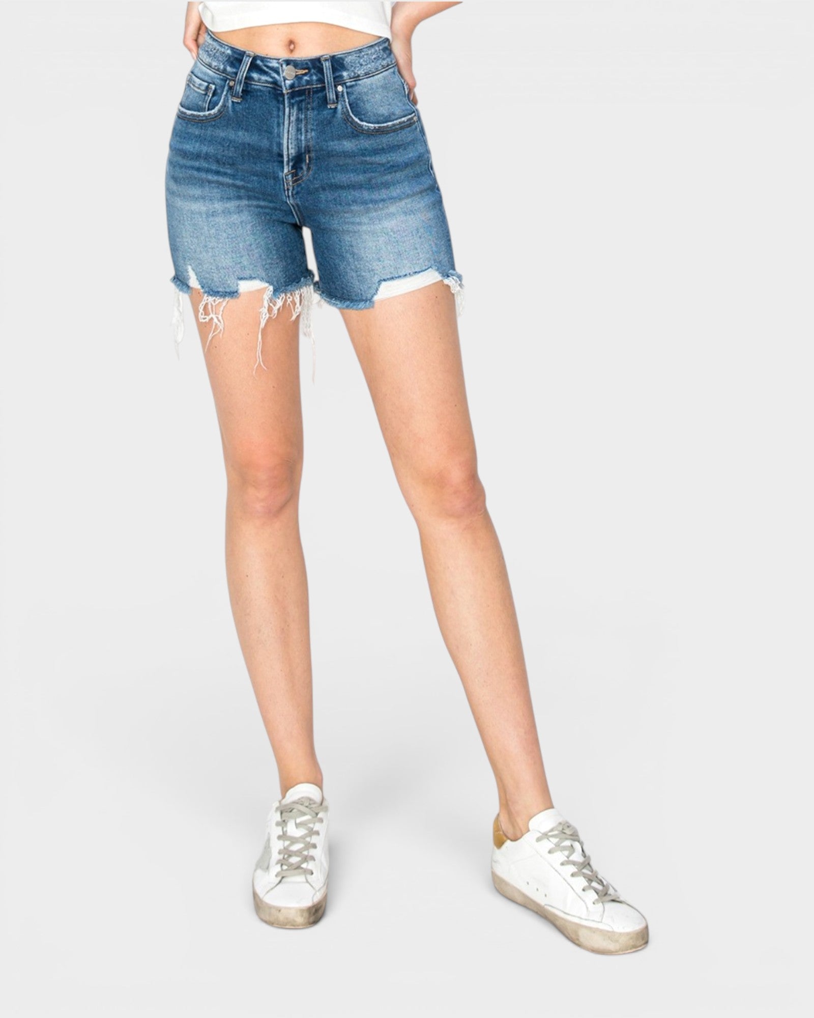 A model wearing medium-wash blue denim shorts with a raw, frayed hem, paired with white sneakers, shown from the waist down on a light gray background.