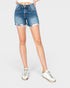 A model wearing medium-wash blue denim shorts with a raw, frayed hem, paired with white sneakers, shown from the waist down on a light gray background.