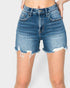 A person wearing medium-wash blue denim shorts with a high-rise fit, frayed raw hems, and distressed detailing, shown from the waist to mid-thigh against a light gray studio background.