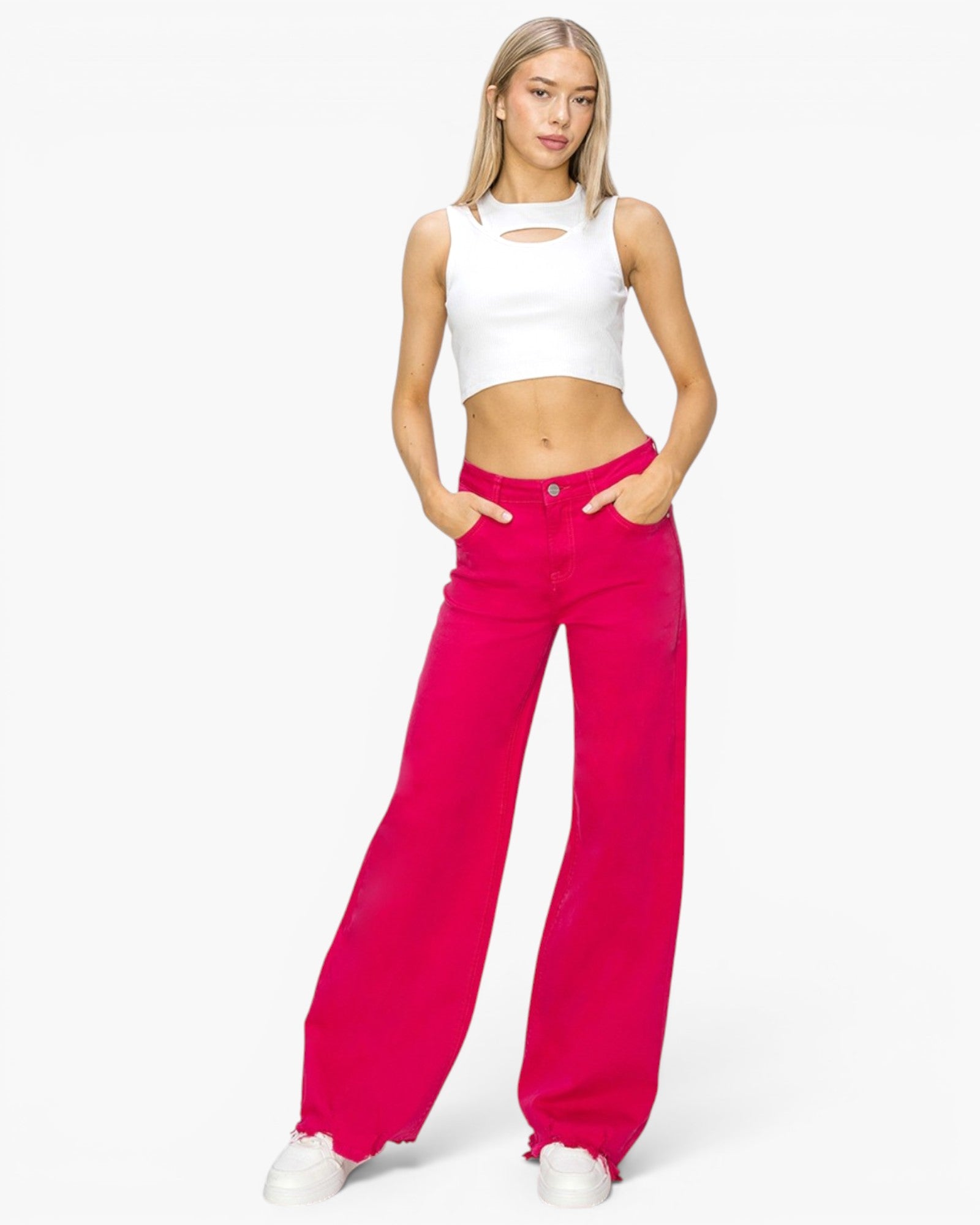 A woman modeling bright fuchsia wide-leg jeans with a white cropped tank top featuring a chest cutout, standing against a plain white studio background.