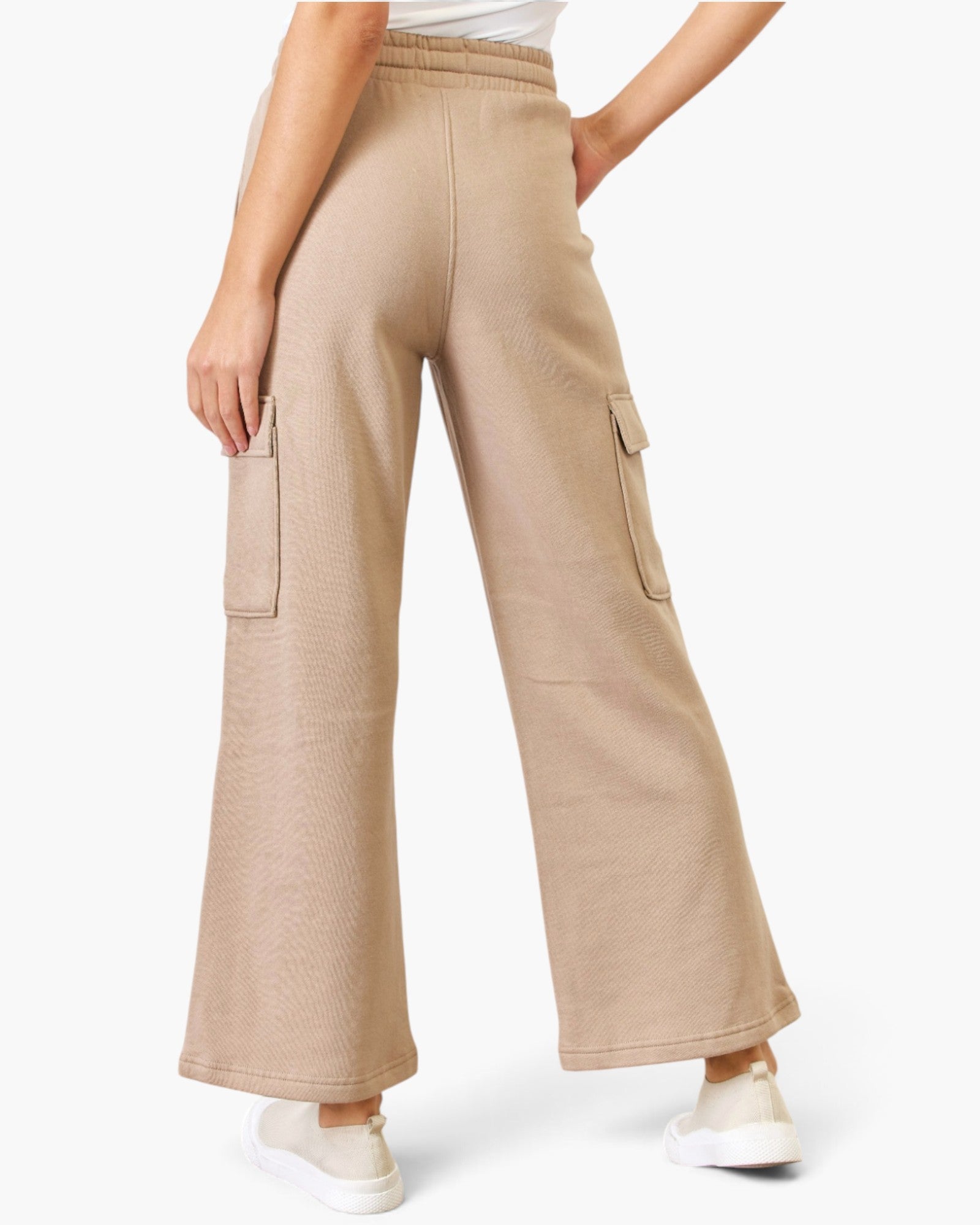 A model is shown from the waist down wearing beige wide-leg cargo pants with side flap pockets, paired with white sneakers against a light gray studio background.