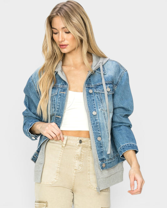 Woman wearing a denim jacket over a gray hoodie with a white top and beige pants on a light gray background