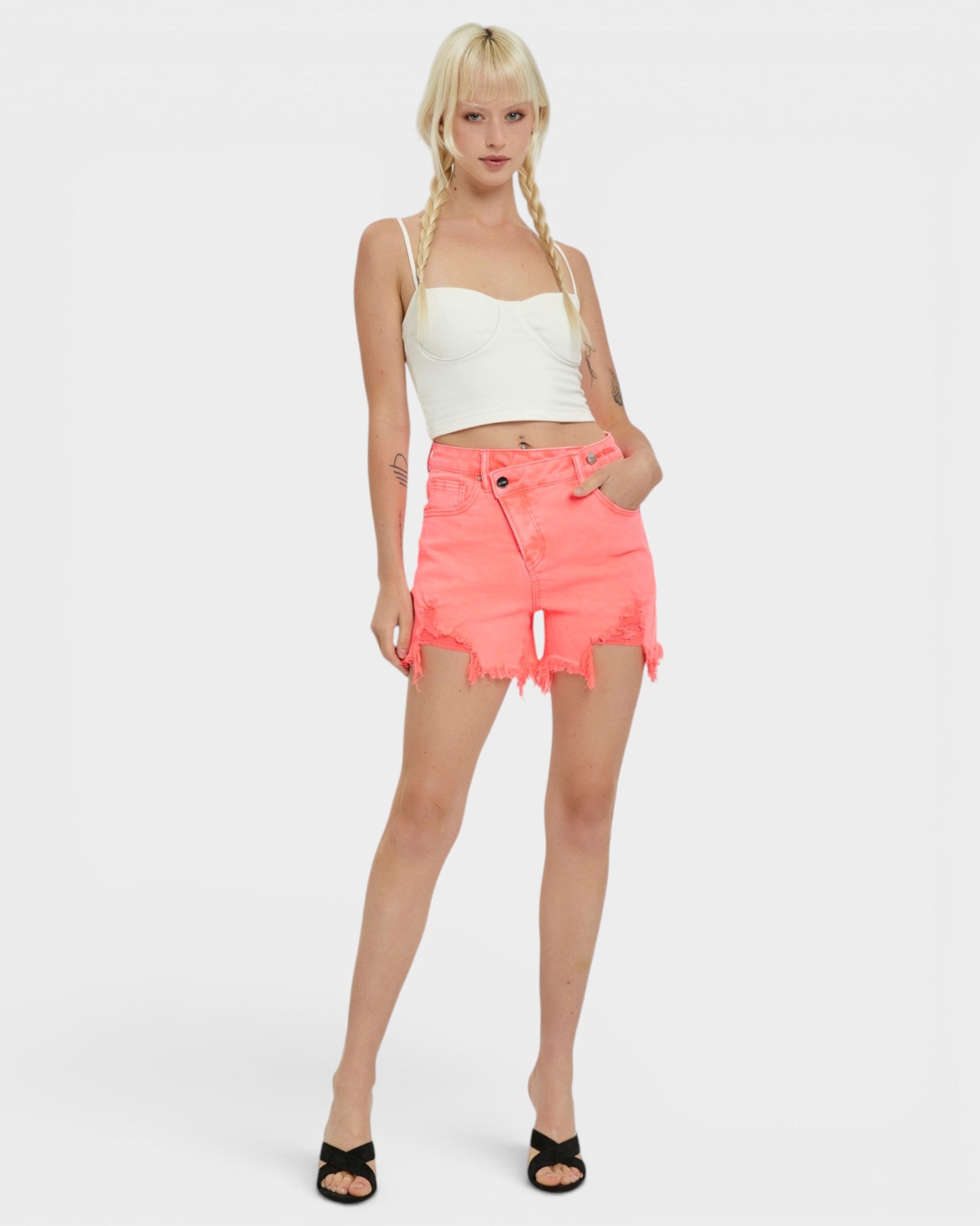 A blonde model with braids stands against a light gray studio background wearing a white cropped camisole, bright coral-pink frayed denim shorts, and black slide sandals.