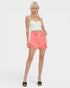 A blonde model with braids stands against a light gray studio background wearing a white cropped camisole, bright coral-pink frayed denim shorts, and black slide sandals.