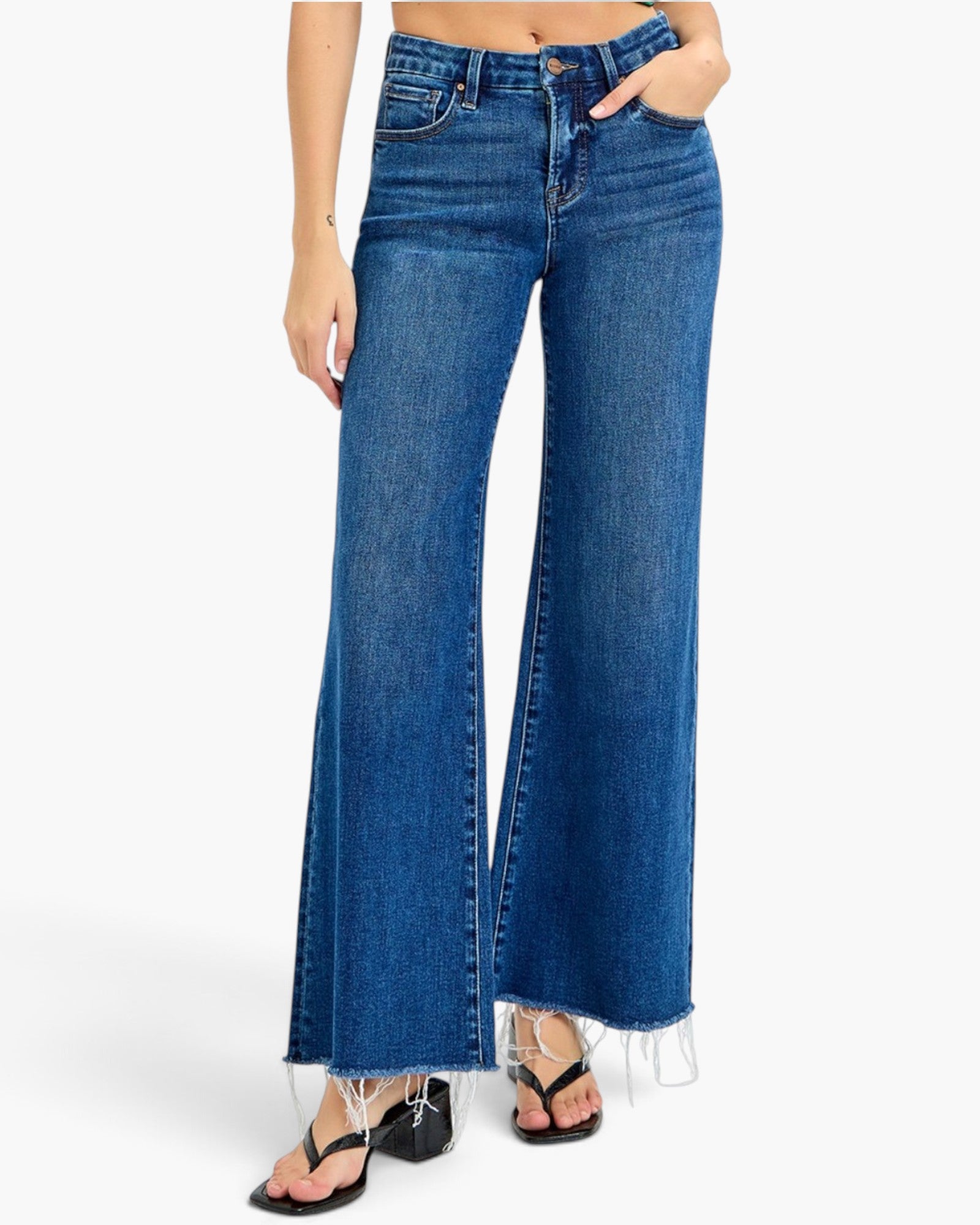 A model wearing high-waisted, wide-leg blue denim jeans with a raw frayed hem, styled with black strappy heels against a plain white background.
