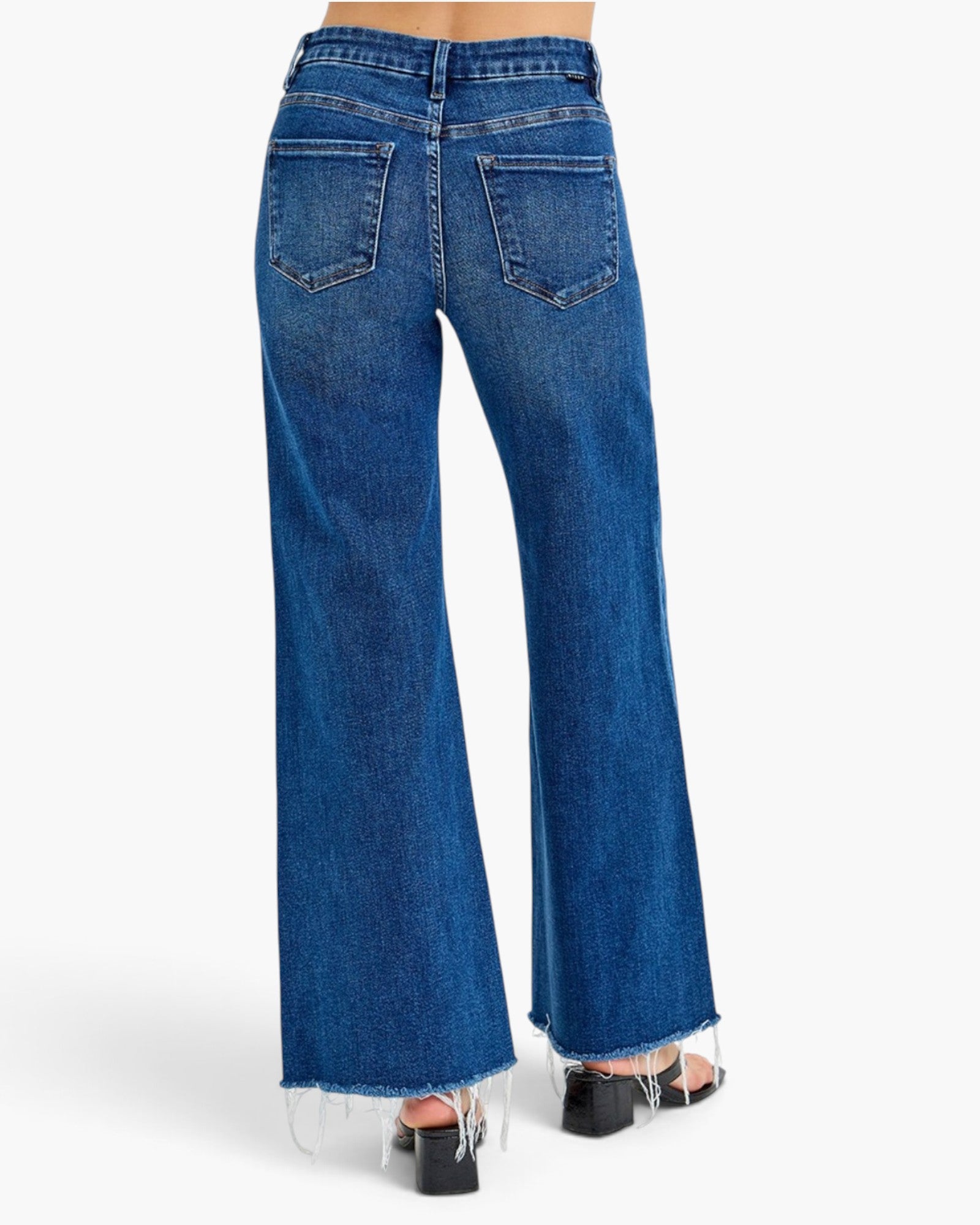 Rear view of a person wearing blue wide-leg denim jeans with a frayed hem and black block-heel sandals against a light gray studio background.