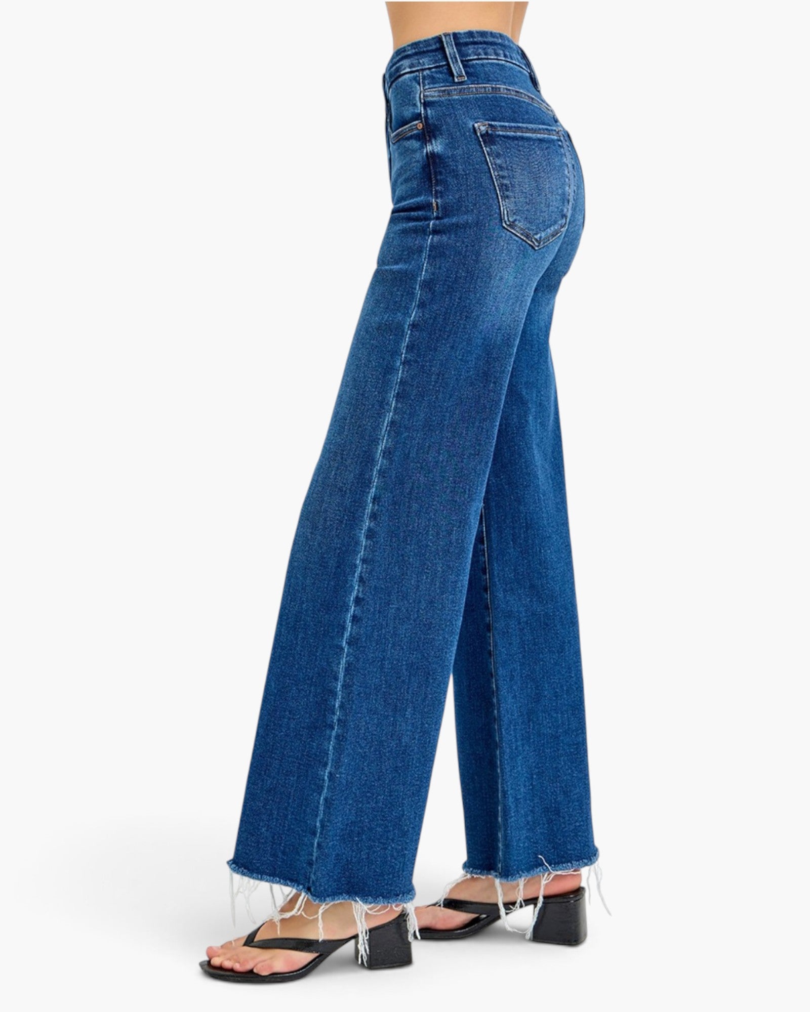 A model shown from the waist down wearing high-waisted, wide-leg blue denim jeans with raw frayed hems and black block-heel sandals, photographed against a clean white background.