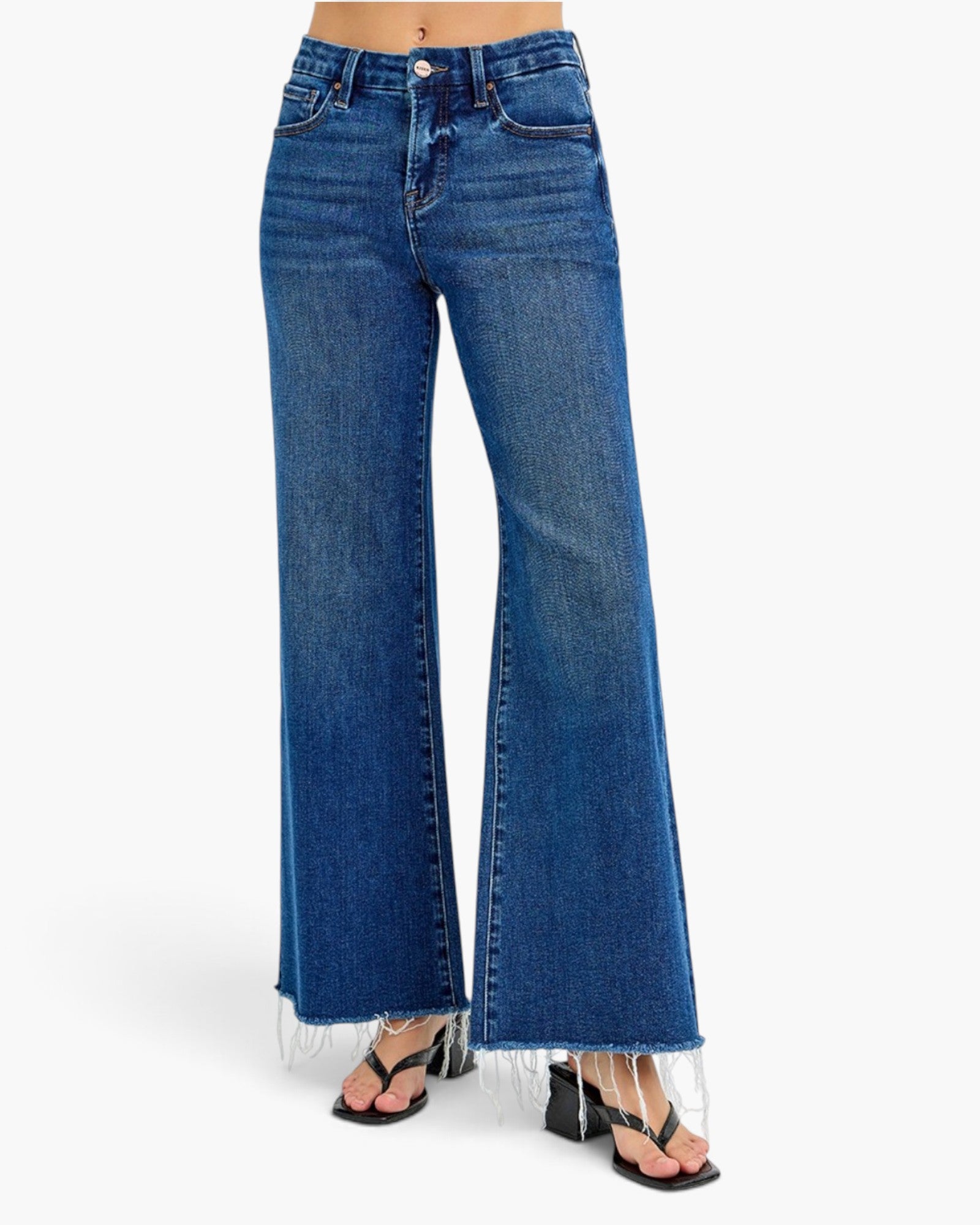 A model wearing a pair of medium-wash blue denim flare jeans with a raw frayed hem, paired with black sandals, shown against a plain white studio background.