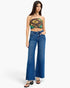 A model wearing a strapless tube top with a colorful baroque pattern and high-waisted, wide-leg blue denim jeans, standing against a plain white studio background.