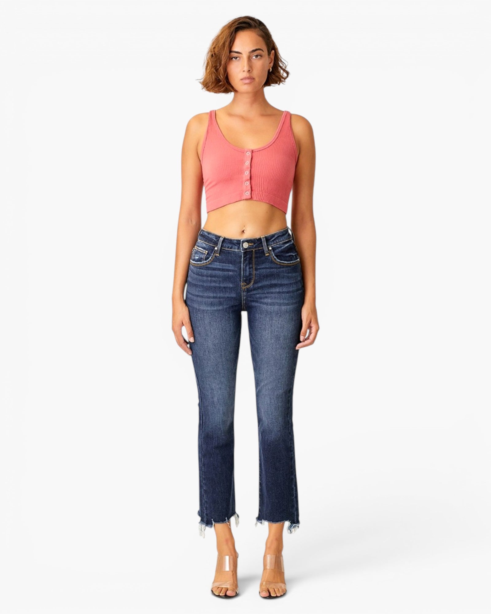 A woman modeling a coral pink ribbed crop top with a front button placket and dark blue cropped denim jeans with a raw hem, standing against a light gray studio background.
