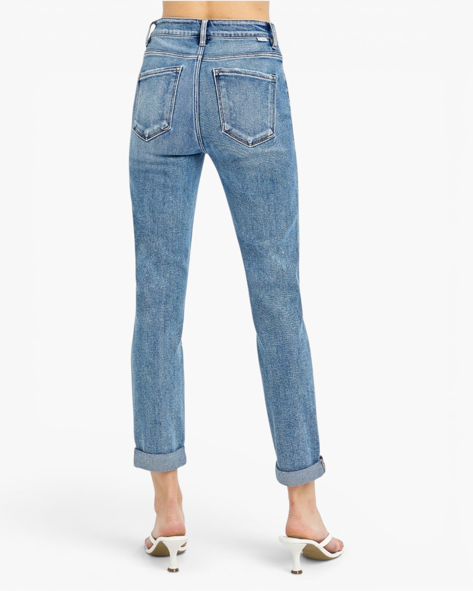 Rear view of a person wearing light blue denim jeans with rolled cuffs and white heeled sandals against a plain white studio background.