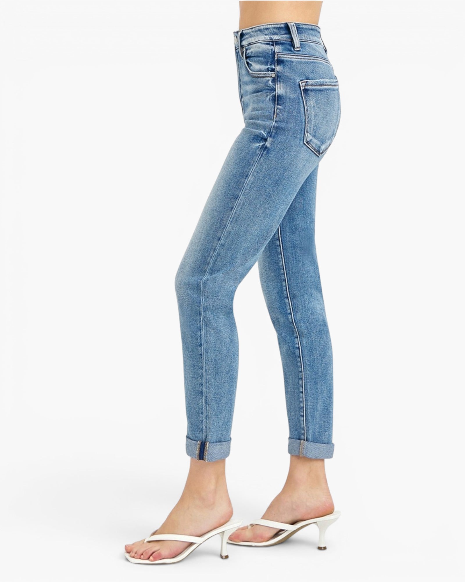 A side profile view of a model wearing light blue high-waisted skinny jeans with rolled cuffs, paired with white heeled thong sandals, against a plain white background.