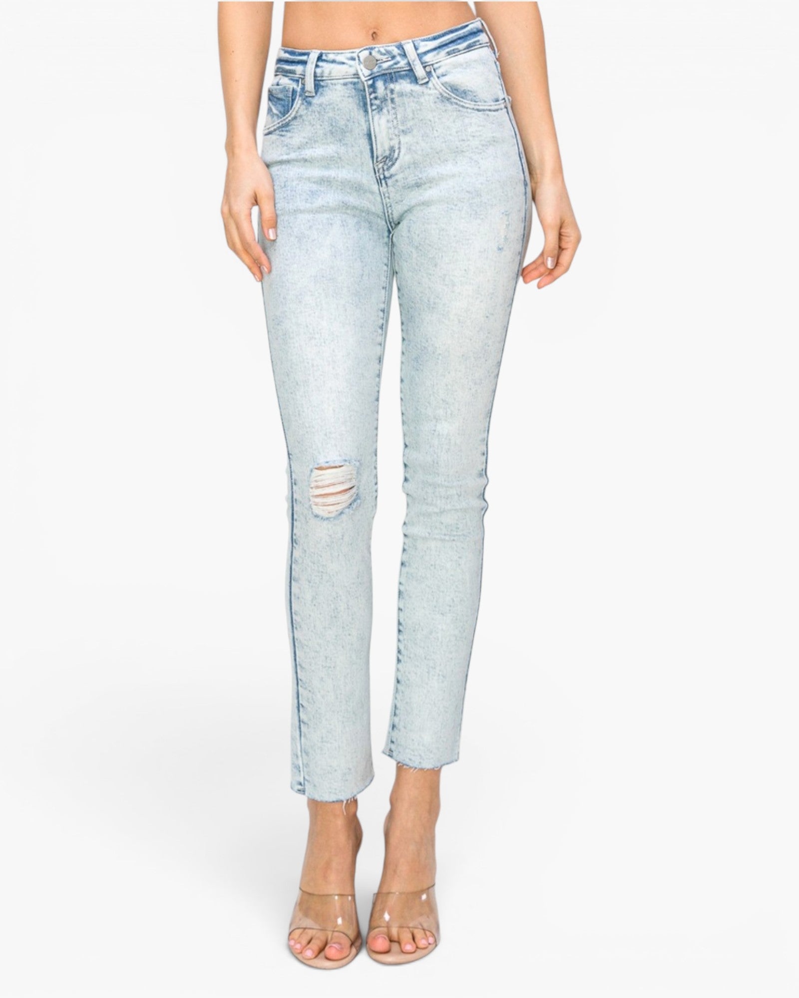 A model wearing light-wash blue distressed skinny jeans with a cropped ankle length, paired with clear high-heeled sandals, against a plain white background.