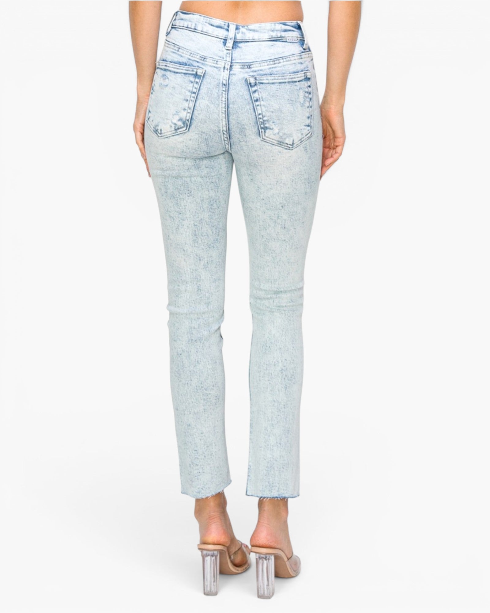 Back view of a model wearing light-wash blue denim jeans with a raw hem and clear-strap high-heeled sandals against a white studio background.