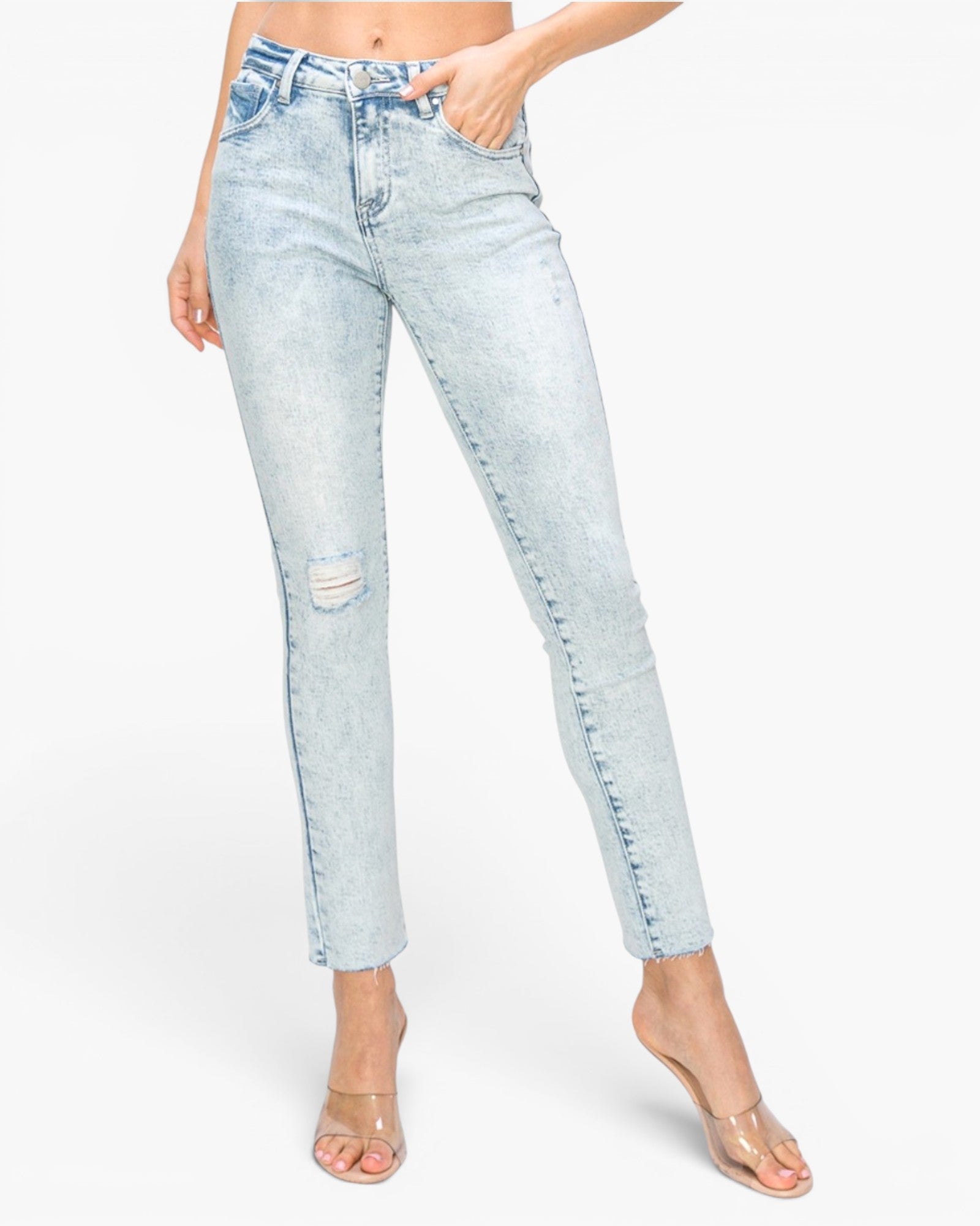 A model wearing light-wash blue distressed skinny jeans with a cropped ankle length, paired with clear strap heels, against a plain white studio background.
