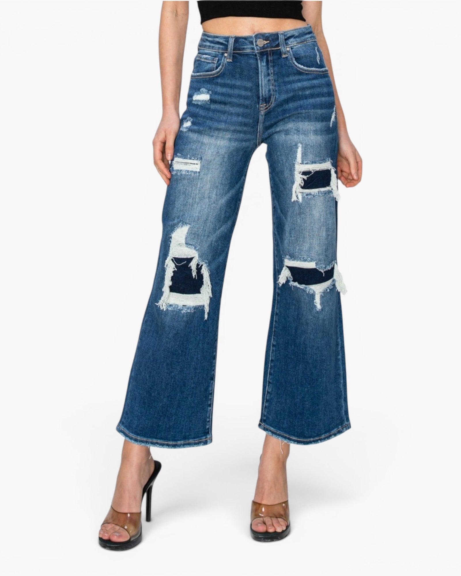 A model wearing a pair of high-waisted, wide-leg cropped blue denim jeans with distressed rips and frayed details, paired with brown heeled sandals against a light gray background.
