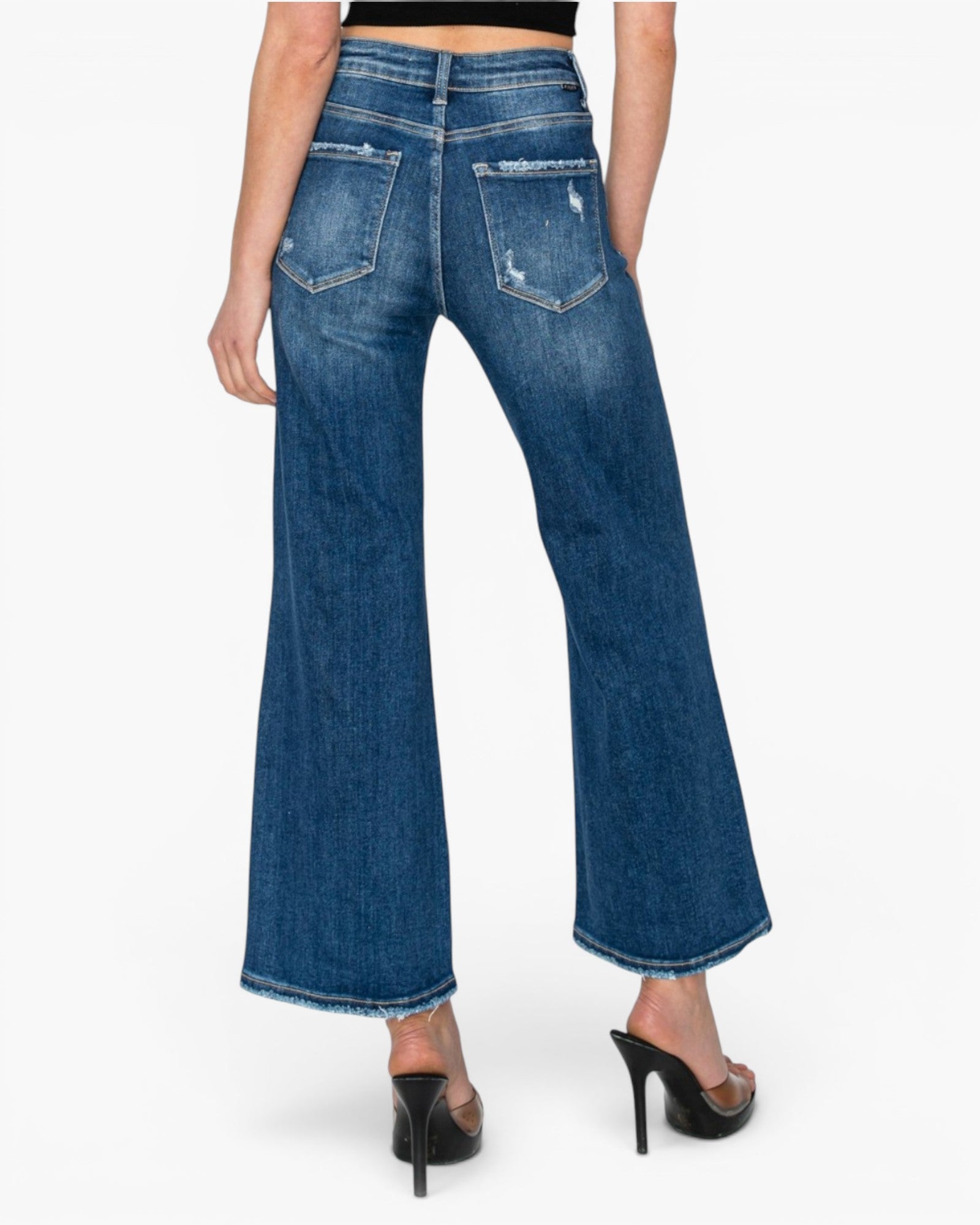 Rear view of a model wearing blue wide-leg cropped jeans with distressed details and frayed hems, paired with black high-heeled sandals against a light gray background.