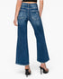 Rear view of a model wearing blue wide-leg cropped jeans with distressed details and frayed hems, paired with black high-heeled sandals against a light gray background.
