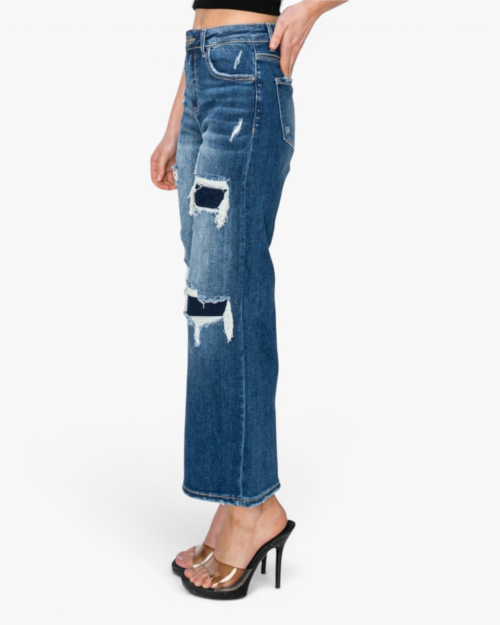 A side view of a model wearing blue distressed denim jeans with ripped details and frayed edges, paired with black high-heeled sandals against a plain white background.