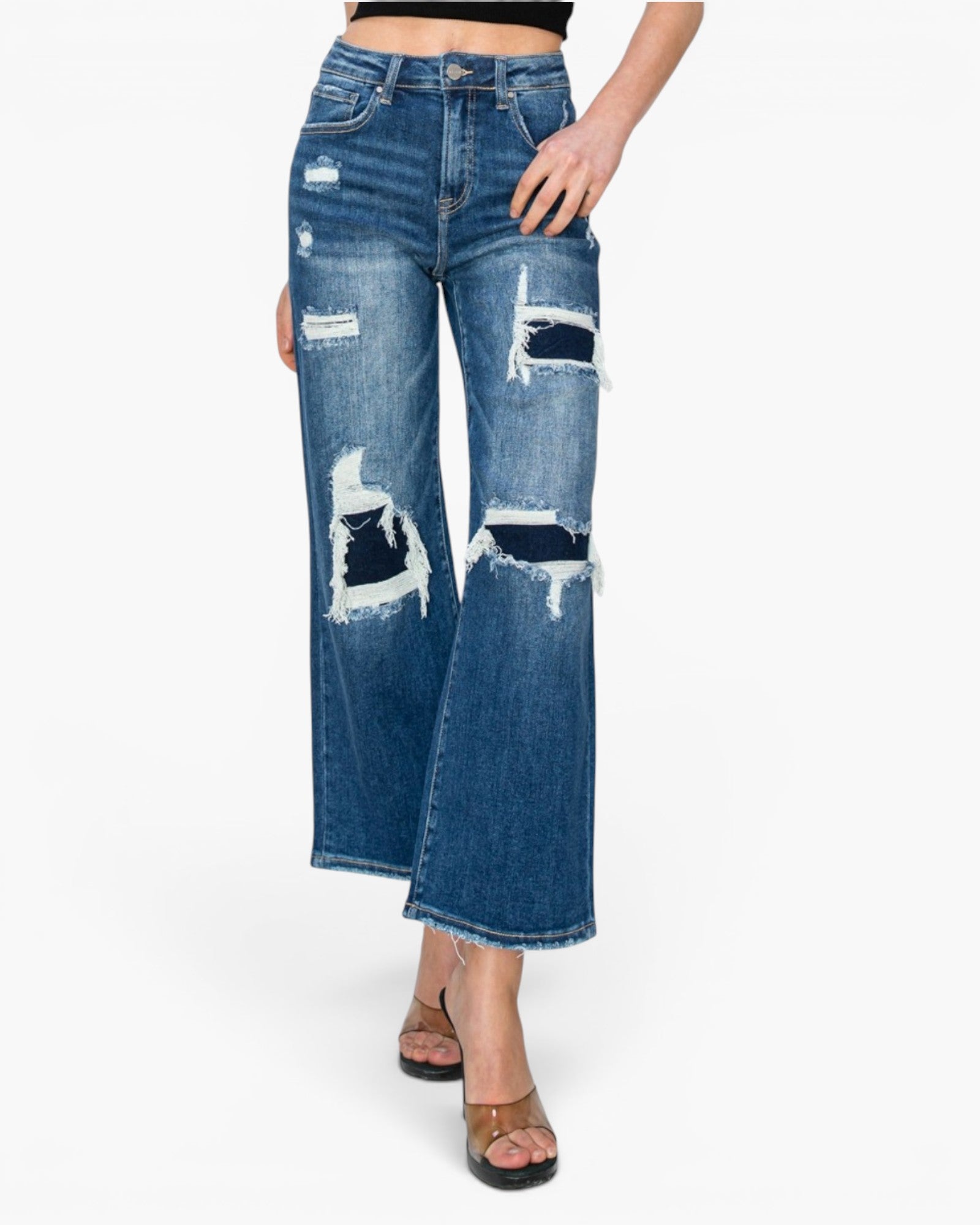 A model wearing a pair of medium-wash blue denim jeans with distressed rips and frayed details on the thighs and knees, paired with brown heeled sandals against a plain white background.
