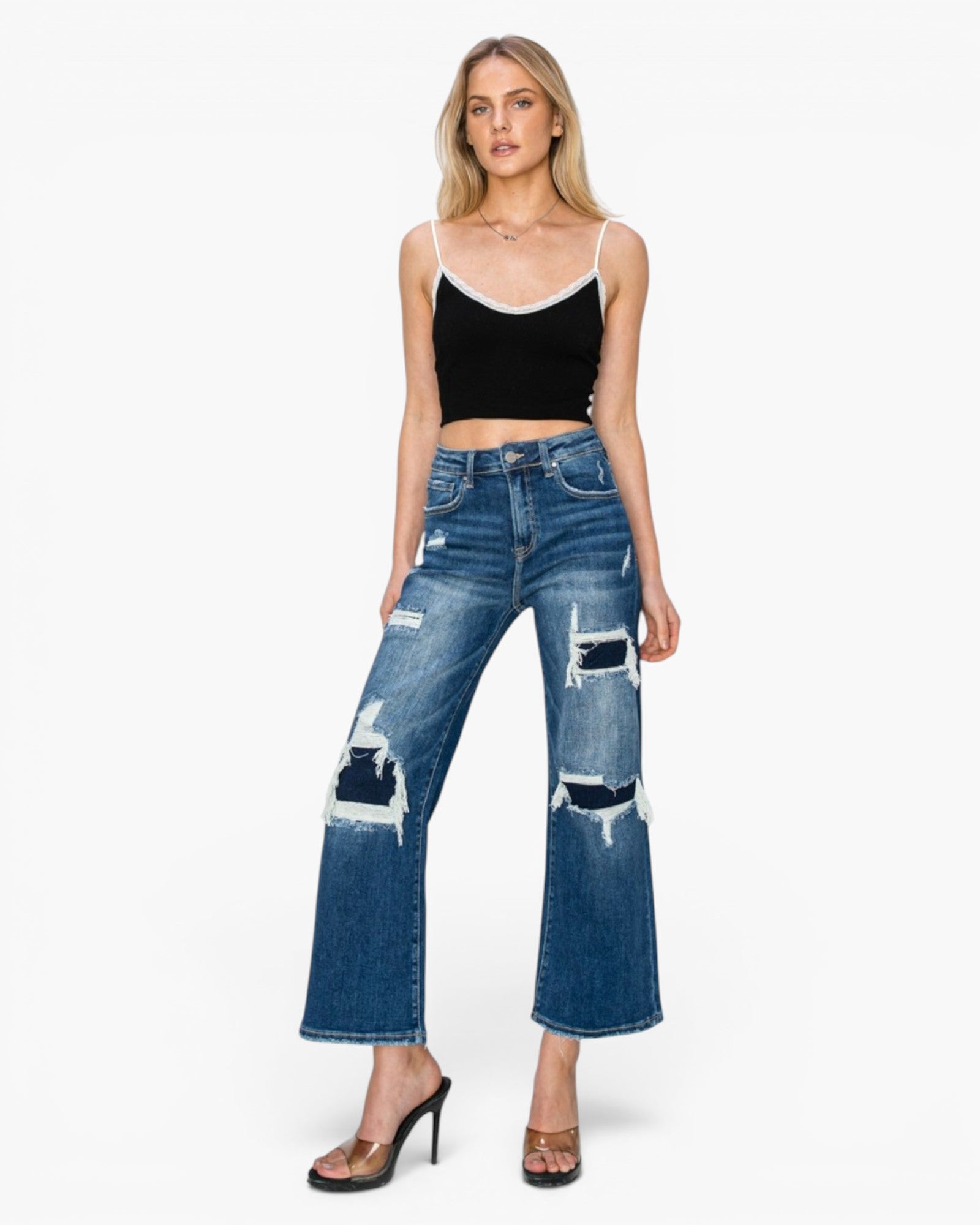 A model wearing a pair of high-waisted, wide-leg blue jeans with distressed rips and frayed details, styled with a black camisole top and brown heeled sandals against a light gray background.