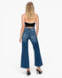 A woman viewed from the back wearing blue wide-leg flare jeans, a black spaghetti-strap crop top, and black high heels against a plain white studio background.