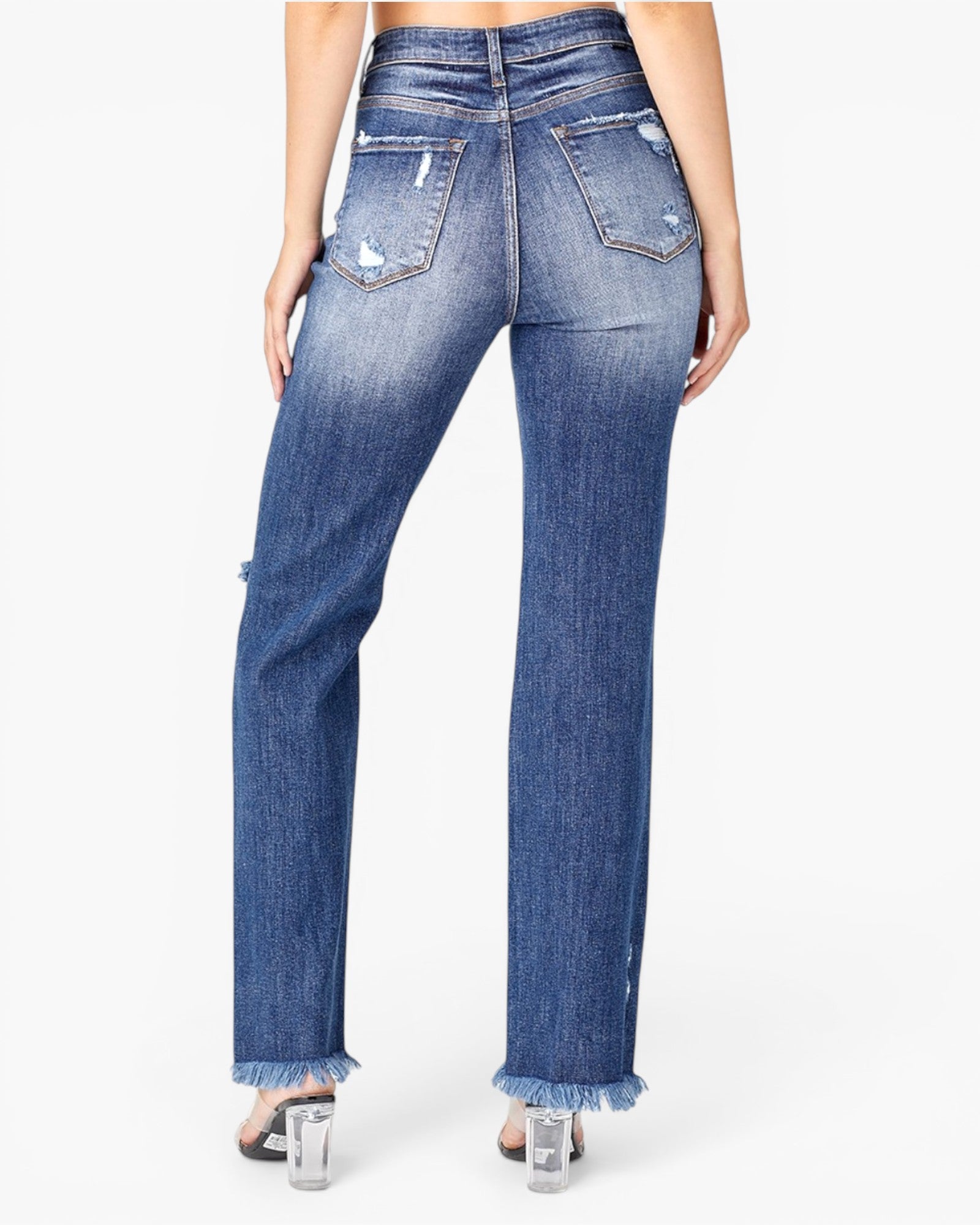 Rear view of a model wearing high-waisted, straight-leg blue denim jeans with distressed details and frayed hems, paired with clear high-heeled sandals against a white studio background.