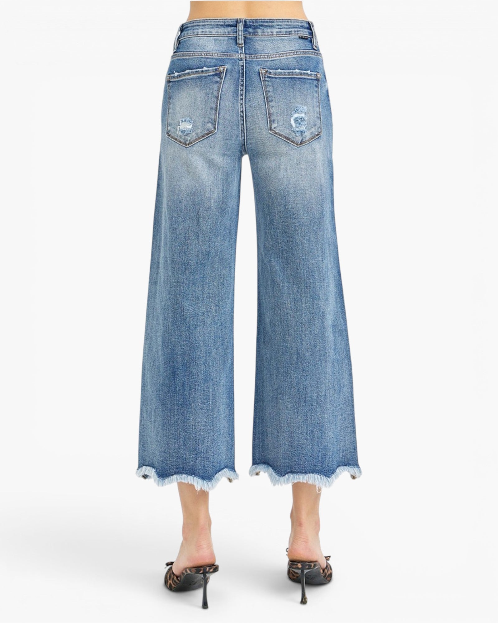 Back view of a model wearing light blue wide-leg cropped denim jeans with a frayed hem and distressed details on the back pockets, paired with black pointed-toe mule heels.