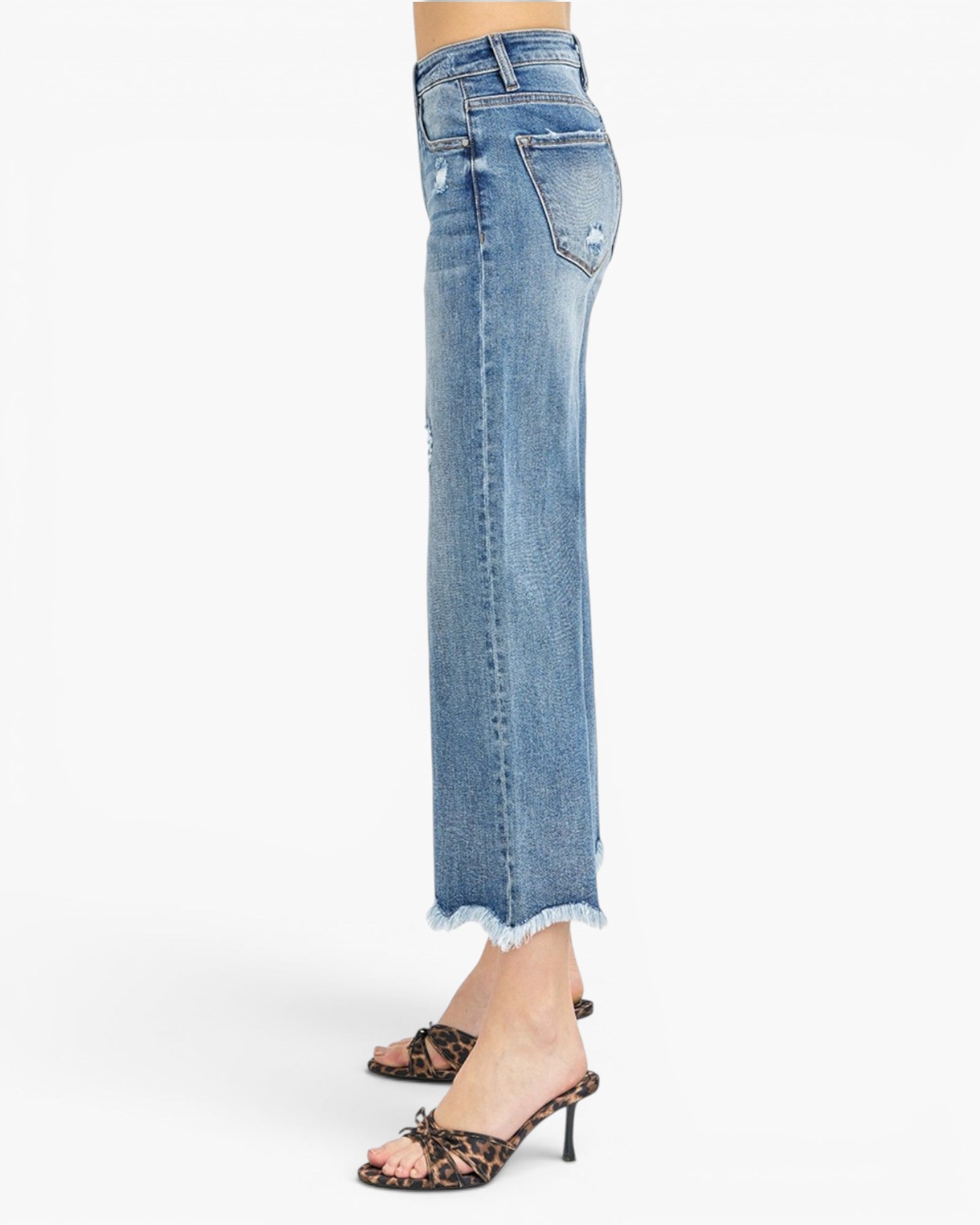 A person shown from the waist down wearing light-wash blue cropped denim jeans with a frayed hem and distressed details, paired with leopard-print heeled sandals against a plain white studio background.