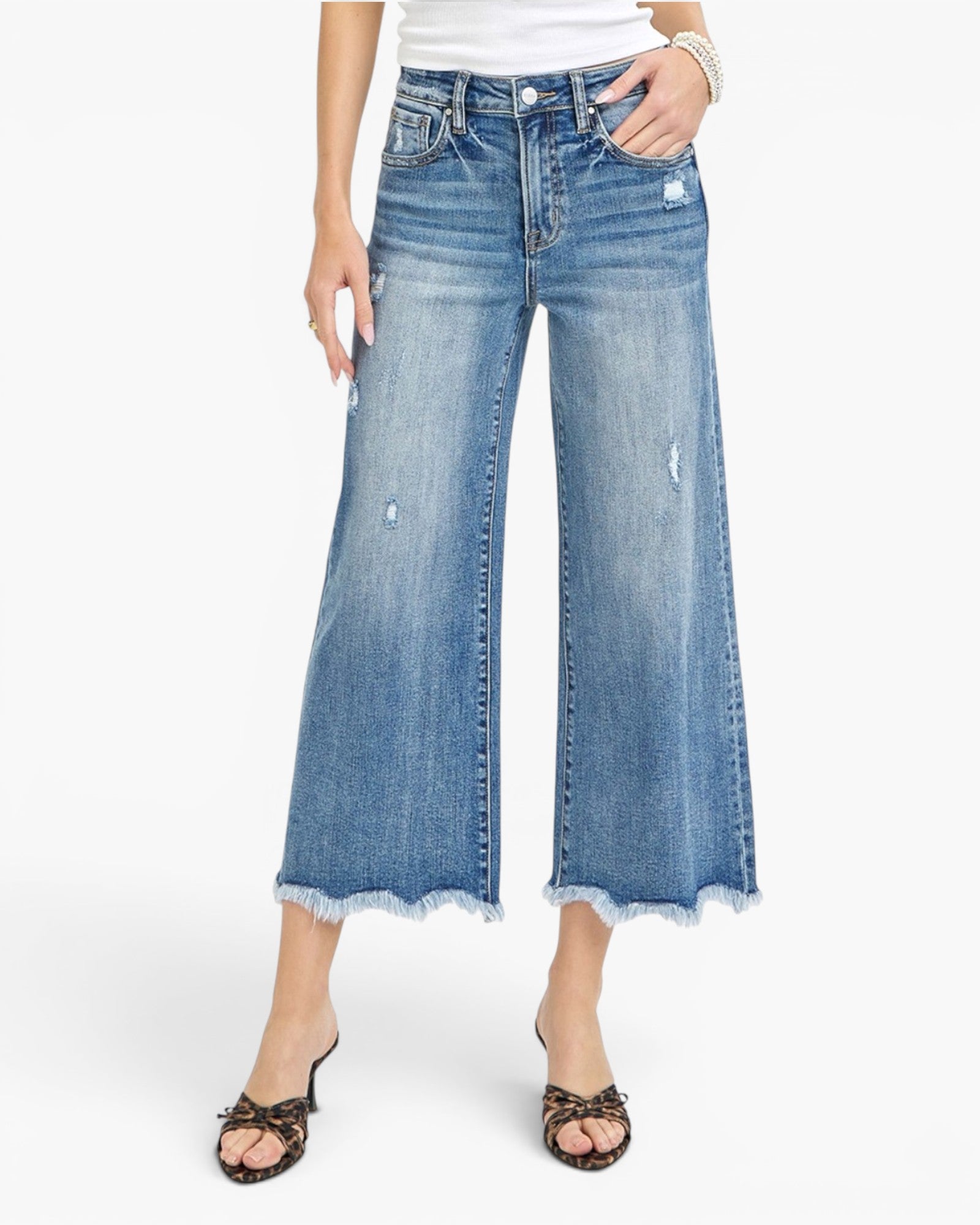 A model wearing a pair of wide-leg cropped blue denim jeans with distressed details and frayed hems, paired with leopard-print heeled sandals against a plain white background.