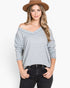 A woman wearing a light gray off-the-shoulder long-sleeve knit top, black jeans, and a brown wide-brim hat, standing against a plain white studio background.