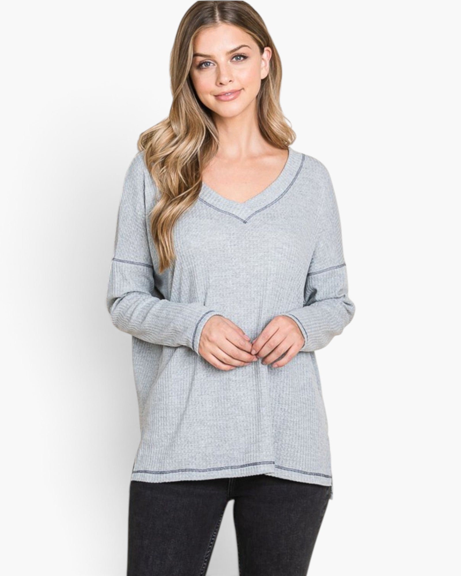 A woman modeling a light gray long-sleeve V-neck knit top with subtle contrast stitching, paired with black pants, against a plain white studio background.