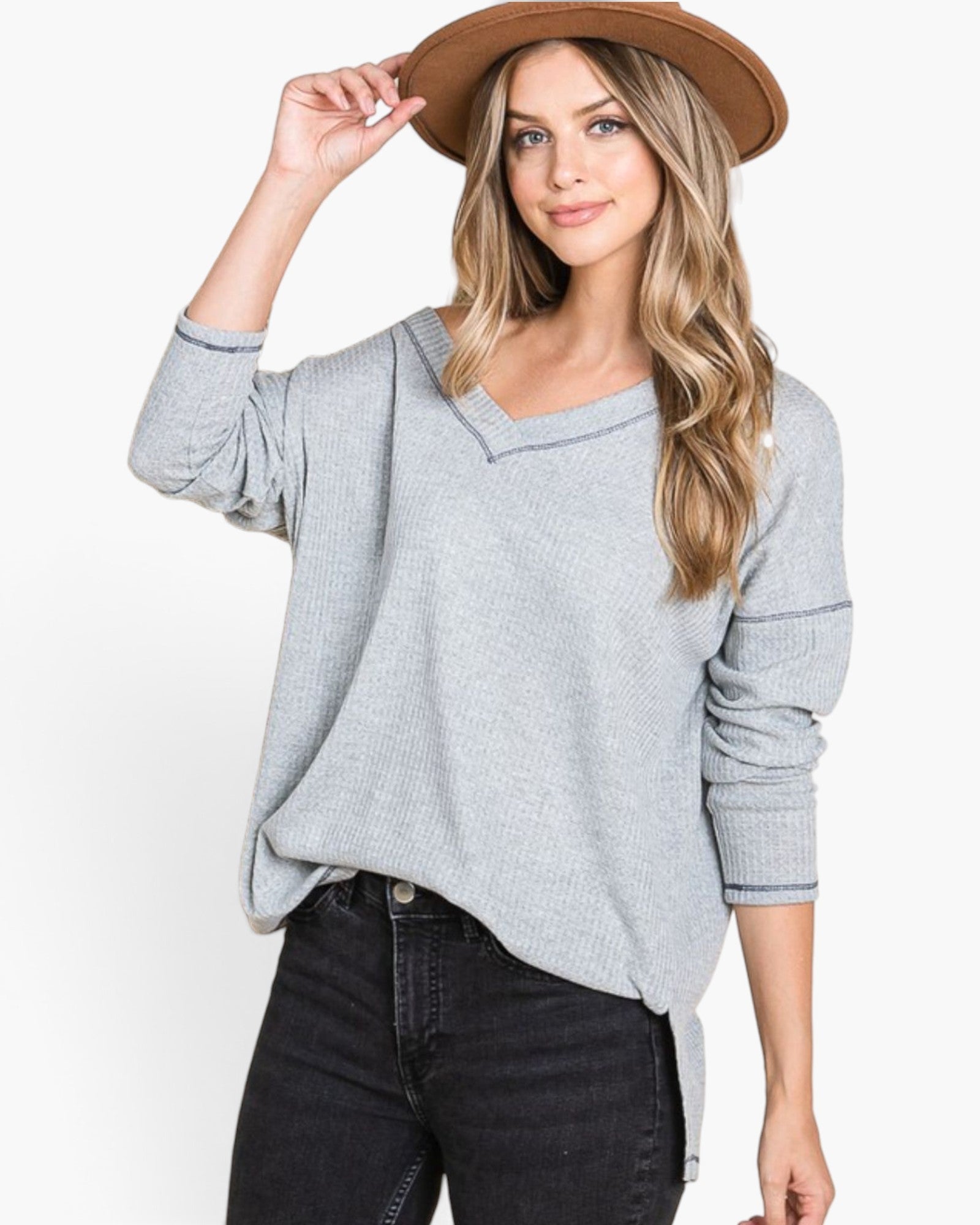 A woman wearing a light gray waffle-knit sweater with a V-neck and long sleeves, paired with black jeans and a brown wide-brim hat, against a plain white background.