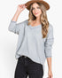 A woman wearing a light gray waffle-knit sweater with a V-neck and long sleeves, paired with black jeans and a brown wide-brim hat, against a plain white background.