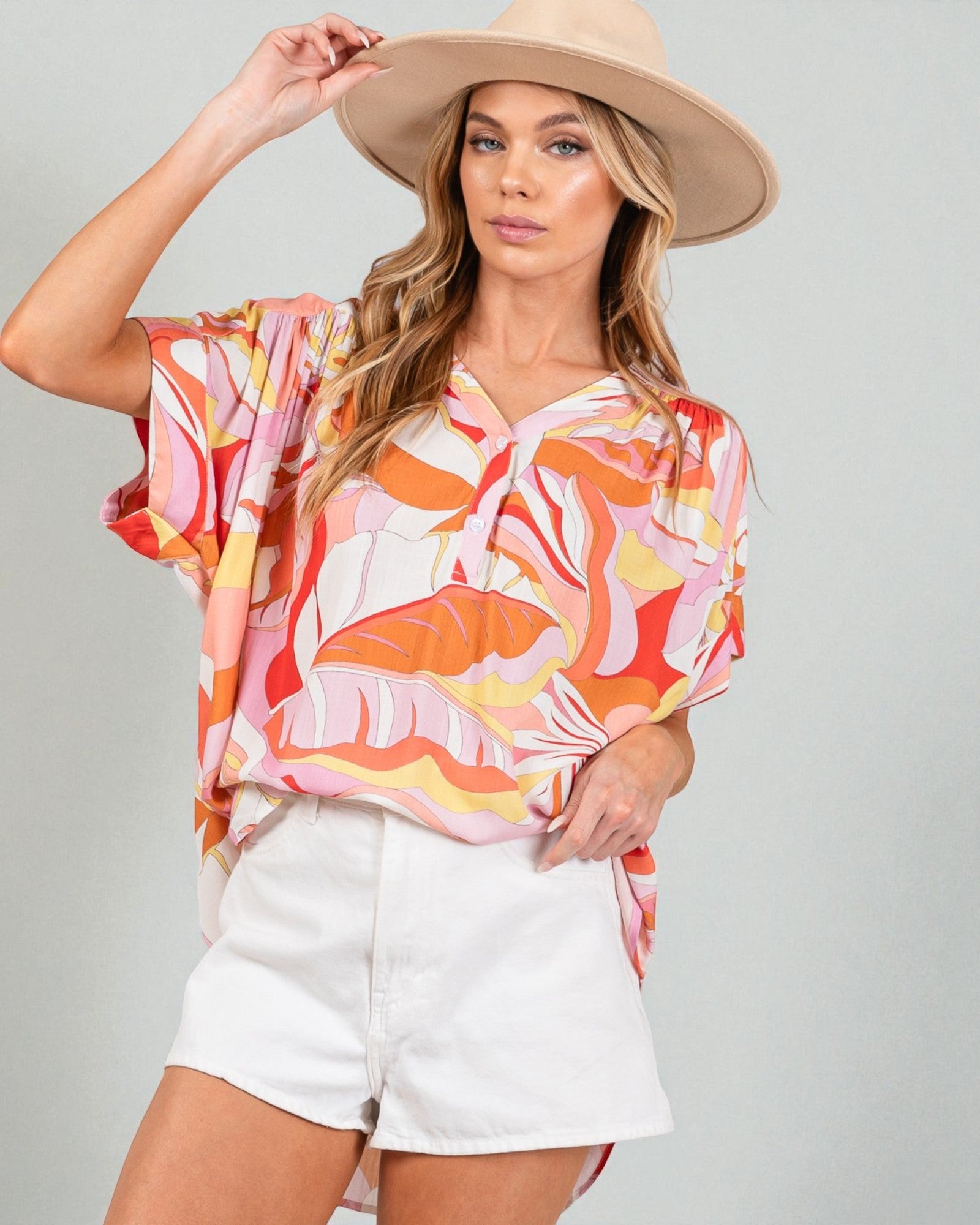 Woman wearing a colorful blouse and white shorts with a beige hat on a gray background