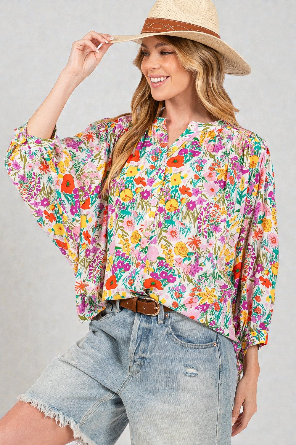 Woman wearing a colorful floral blouse and denim shorts with a straw hat on a gray background