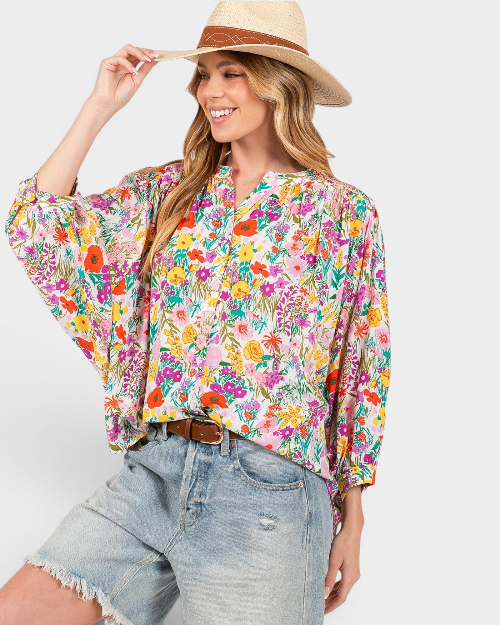 A woman wearing a vibrant, multicolor floral print blouse with balloon sleeves, styled with light-wash denim shorts and a straw hat against a solid light gray background.
