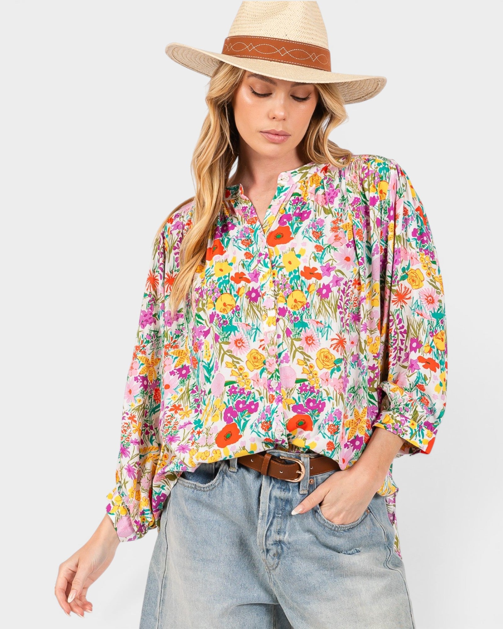 A woman wearing a long-sleeve button-up blouse with a vibrant multicolor floral print, styled with light-wash blue jeans, a brown belt, and a straw hat with a brown band, set against a plain light gray background.