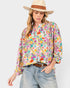 A woman wearing a long-sleeve button-up blouse with a vibrant multicolor floral print, styled with light-wash blue jeans, a brown belt, and a straw hat with a brown band, set against a plain light gray background.